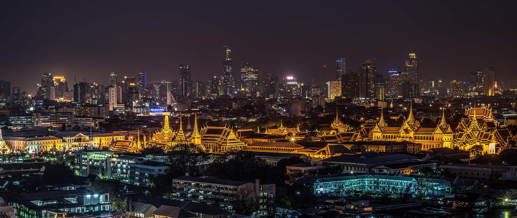 Skyline of Bangkok, Thailand at night, with low buildings in the foreground, a temple complex in the center, and modern skyscrapers in the background