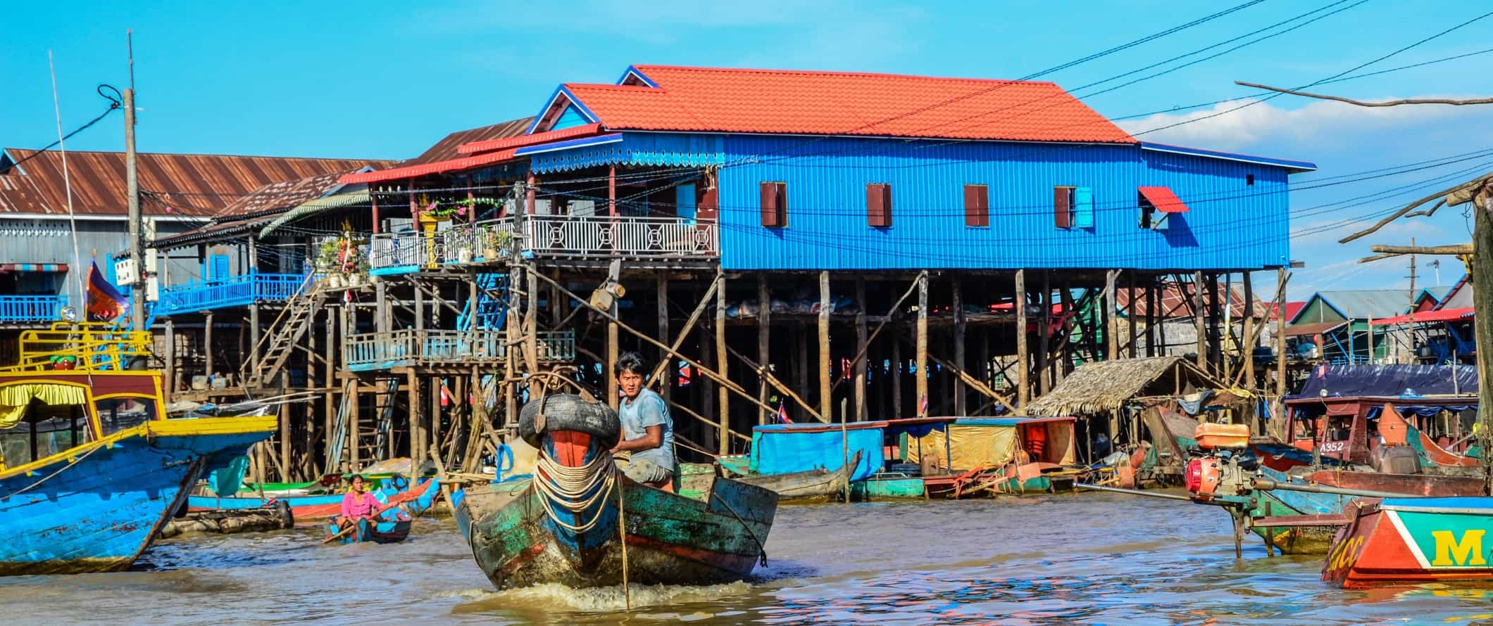 Man driving a boat down a waterway in front of brightly colored houses on stilts in Tonle Sap, Cambodia