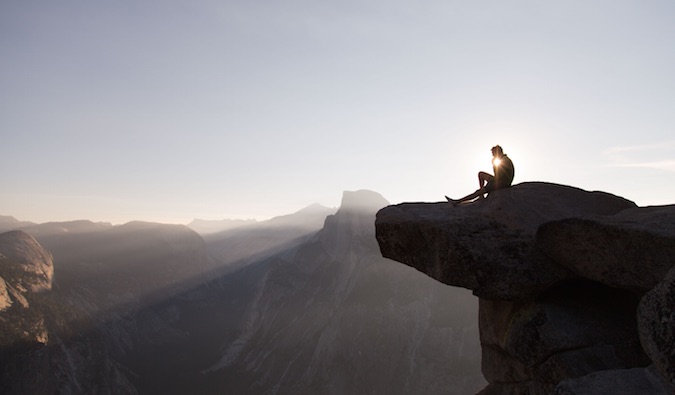 A solo traveler sitting on the edge of a cliff thinking during the sunset