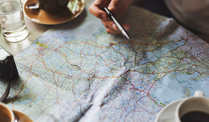 a man's hand holding a pen pointing towards a place on a map, camera, coffee cups, and a glass of water